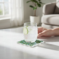 A hand reaches for sparkling water with ice and cucumber slices, set atop The Darling Effect's Everbloom Forest Green Table Talk Acrylic Coaster Set on a white table in a sunlit room with a plant and sofa in the background.
