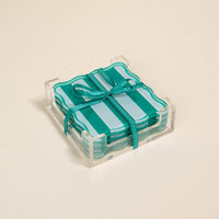 The Darling Effect Sicily Stripes Teal Table Talk Acrylic Coaster Set features heat-resistant, teal and turquoise striped coasters with scalloped edges in a clear holder, tied with a teal ribbon—ideal for chic table décor.