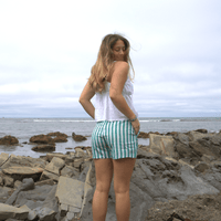 A woman with long hair stands on rocky terrain by the ocean, wearing a white sleeveless top and The Darling Effect's Sicily Stripes Teal All Day Boxer Shorts, made from breathable fabric, as she looks over her shoulder on a cloudy day.