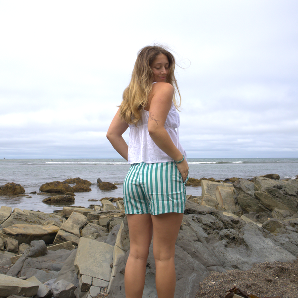 A woman with long hair stands on rocky terrain by the ocean, wearing a white sleeveless top and The Darling Effect's Sicily Stripes Teal All Day Boxer Shorts, made from breathable fabric, as she looks over her shoulder on a cloudy day.