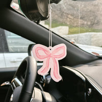 The Bow Pink Sweet Ride Car Air Freshener by the-darling-effect hangs from your rearview mirror, adding a unique scent and a stylish touch to your car’s interior.