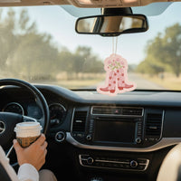 A person holding a coffee cup drives on a sunny day. the-darling-effect’s Cowgirl Boots Sweet Ride Car Air Freshener hangs from the rearview mirror, filling the car with a long-lasting aroma as trees are visible through the windshield.