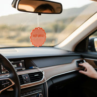 Close-up of a car interior with a hand reaching for the door handle, dashboard screen, and the-darling-effect's Hot Mess Express Sweet Ride Car Air Freshener, delivering long-lasting aroma. Blurred scenery outside completes the vibe.