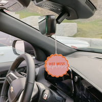 A car interior with steering wheel, dashboard, and rearview mirror features the-darling-effect's Hot Mess Express Sweet Ride Car Air Freshener in orange and white, adding a fun scent to every drive.