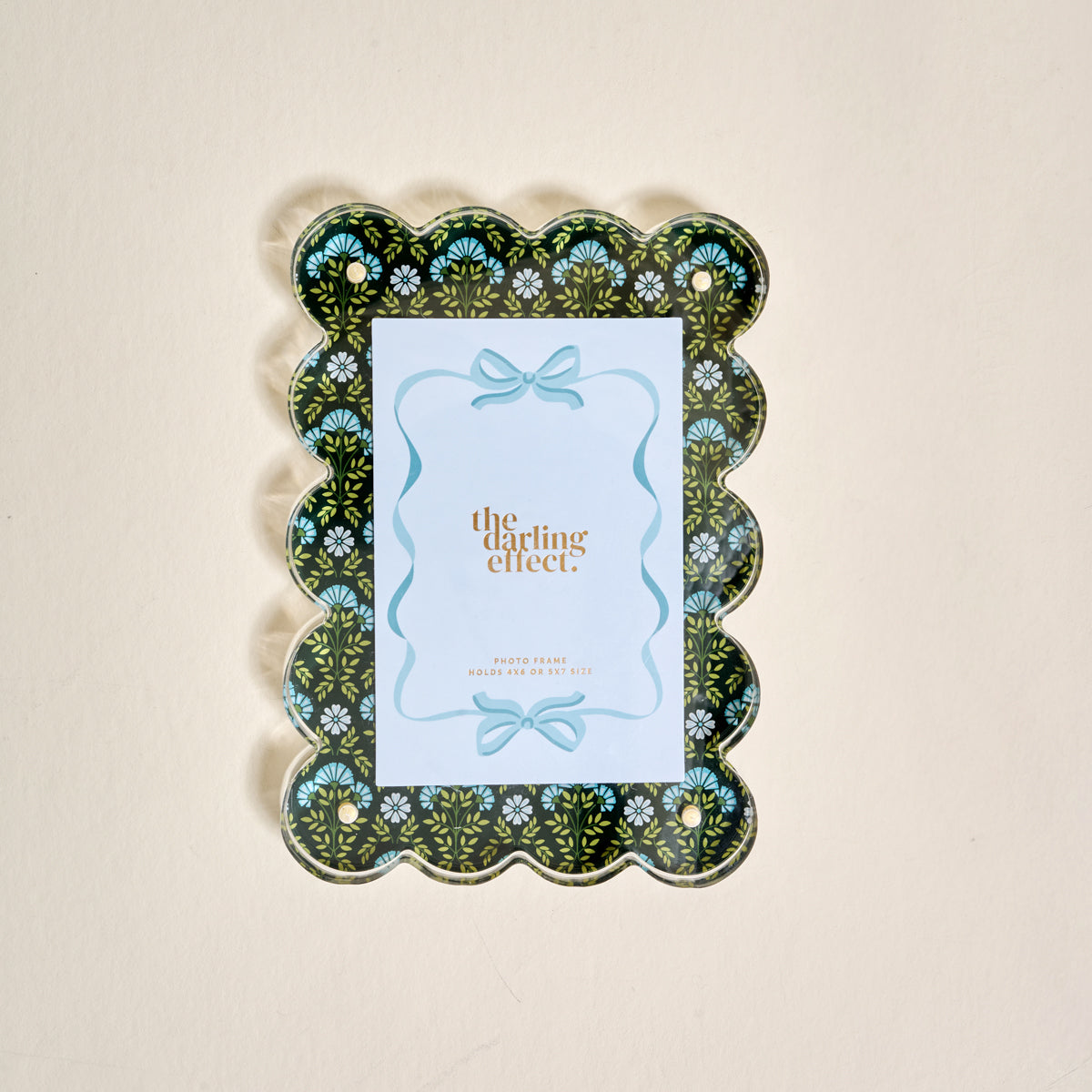 The Everbloom Forest Green Rectangle Scalloped Acrylic Picture Frame by The Darling Effect displays a card with a decorative blue bow, features a black background with light blue floral accents, and sits on a beige surface.