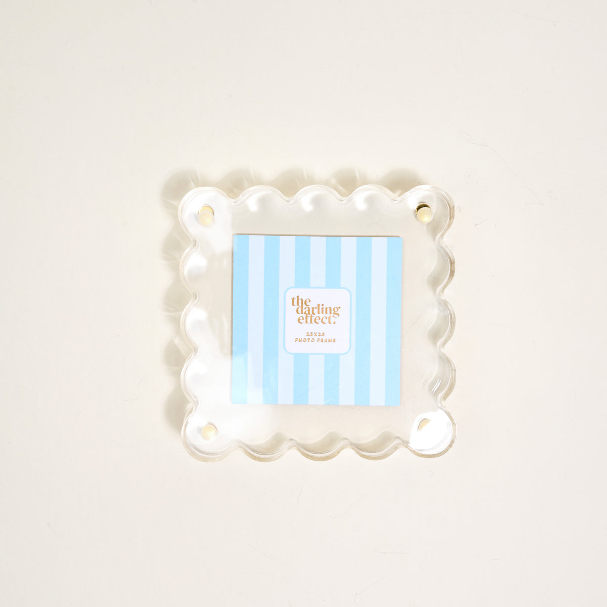 The Darling Effect's Clear Little Moments Acrylic Picture Frame, featuring a square shape, scalloped edges, and four corner holes, displays a blue-and-white striped "the clearing effect" card on a light-colored surface.