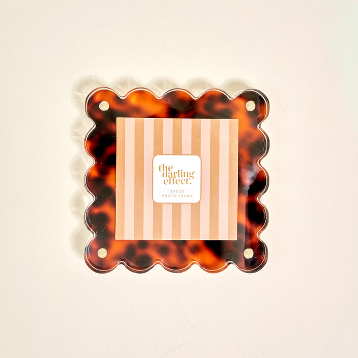 The Darling Effect's Tortoise Little Moments Acrylic Picture Frame features a square scalloped-edge design with a tortoiseshell pattern and corner holes, elegantly displaying a striped card at its center.