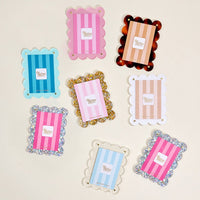 Eight colorful Blush Mini Acrylic Picture Frame Magnets by The Darling Effect are displayed on a light surface. Featuring scalloped edges and "Biscuit Clever" labels, the designs include pink, blue, gold, tortoiseshell, and glitter styles with stripes.