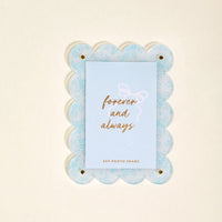 The Darling Effect’s Something Blue Mini Acrylic Picture Frame Magnet features a light blue scalloped design with a floral pattern and displays a card reading "forever and always" in gold script.