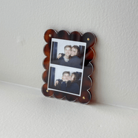 The Darling Effect Tortoise Mini Acrylic Picture Frame Magnet displays two photos—a smiling couple posing together above, and the woman kissing his cheek below—set in a scalloped tortoiseshell frame on a white surface.