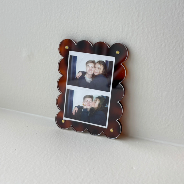 The Darling Effect Tortoise Mini Acrylic Picture Frame Magnet displays two photos—a smiling couple posing together above, and the woman kissing his cheek below—set in a scalloped tortoiseshell frame on a white surface.