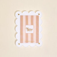 The Darling Effect White Mini Acrylic Picture Frame Magnet features scalloped white edges, gold corner screws, and a pink striped insert with the brand name. Shown against a light beige background.