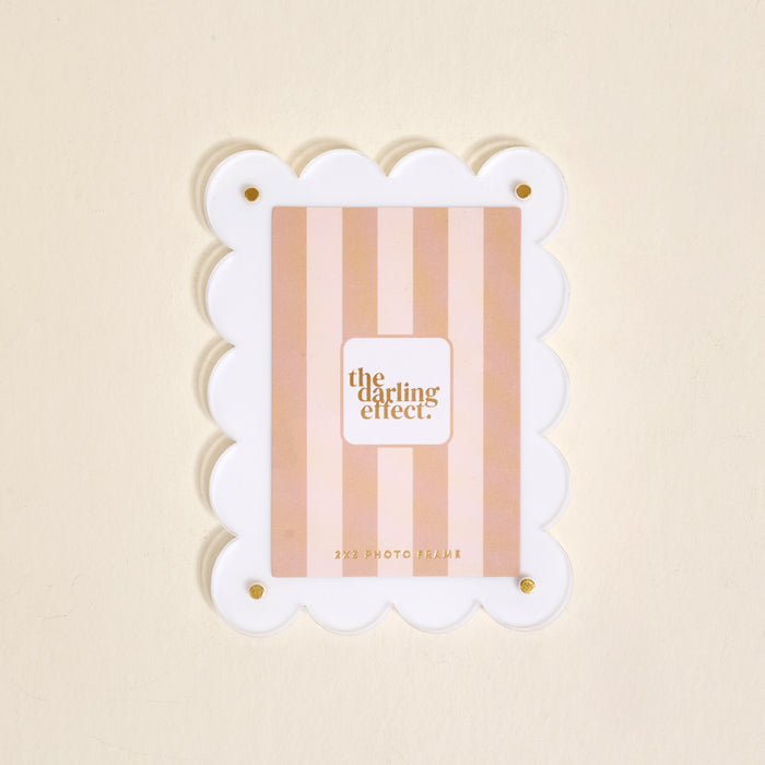 The Darling Effect White Mini Acrylic Picture Frame Magnet features scalloped white edges, gold corner screws, and a pink striped insert with the brand name. Shown against a light beige background.