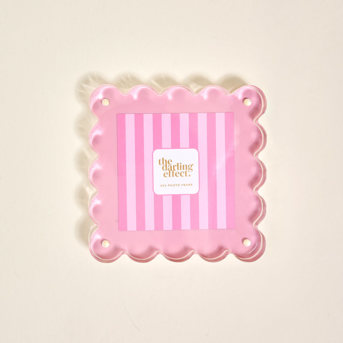 The Darling Effect’s Blush Square Scalloped Acrylic Picture Frame features pink scalloped edges, a striped background, and corner holes for mounting. The modern frame showcases “the darling effect” at its center on a light surface.