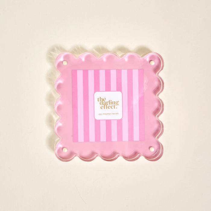 The Darling Effect’s Blush Square Scalloped Acrylic Picture Frame features pink scalloped edges, a striped background, and corner holes for mounting. The modern frame showcases “the darling effect” at its center on a light surface.