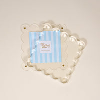 A Clear Square Scalloped Acrylic Frame by the-darling-effect is displayed over a cream background, holding a blue and white striped card with the brand name. This modern 6x6 photo frame features scalloped edges and four corner holes.