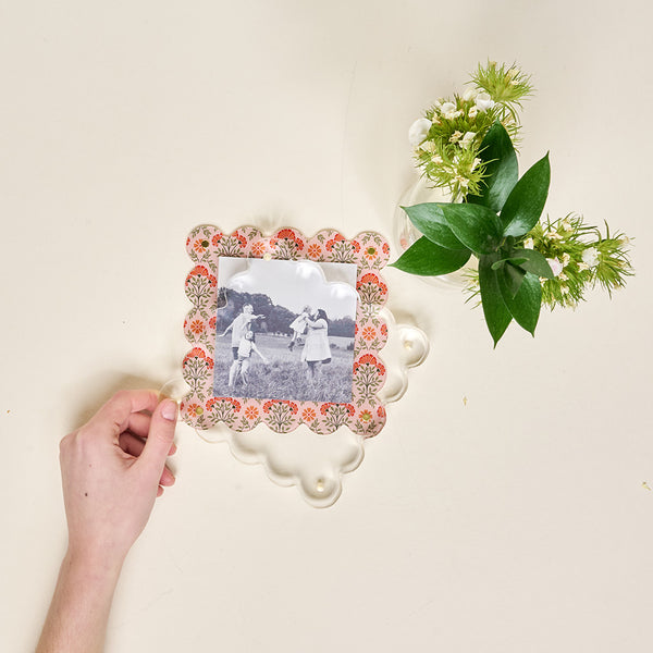 A hand holds The Darling Effect’s Everbloom Dusty Rose Square Scalloped Acrylic Picture Frame displaying a black-and-white photo, set beside a small bouquet of white flowers and greenery on a cream-colored surface.