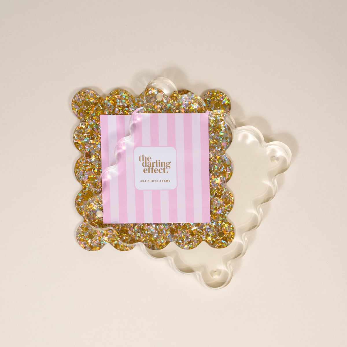 The Darling Effect Gold Glitter Square Scalloped Acrylic Picture Frame, featuring a card with pink and white stripes, sits on a light surface with a wavy shadow.