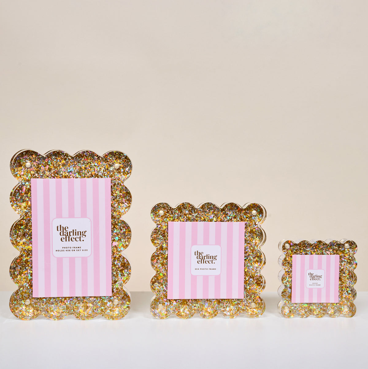 Three gold glitter square scalloped acrylic picture frames by The Darling Effect, each showcasing a pink and white striped print with the brand’s name, displayed against an off-white background.