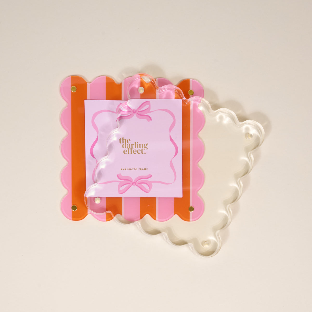 The Darling Effect's Sicily Stripes Pink Square Scalloped Acrylic Picture Frame features pink and orange stripes, gold corner screws, and a clear wavy front for a chic modern look, displayed on a light beige background.