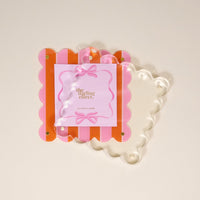 The Darling Effect's Sicily Stripes Pink Square Scalloped Acrylic Picture Frame features pink and orange stripes, gold corner screws, and a clear wavy front for a chic modern look, displayed on a light beige background.