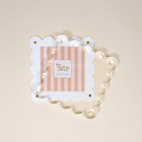 The White Square Scalloped Acrylic Frame by the-darling-effect showcases a pink and white striped card reading "the darling effect." Gold screws accent the clear scalloped frame, which rests on a cream-colored surface.