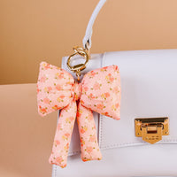 A white handbag with a gold clasp is styled with The Darling Effect’s Peachy Petals Puffy Bow Bag Charm, a coquette-inspired accessory featuring small floral prints and a gold keyring. The background is beige.