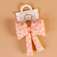 The Darling Effect’s Peachy Petals Puffy Bow Bag Charm features a floral pink bow on a gold keyring, displayed on a white "bagcharm" card against a beige background.