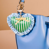 A shiny blue handbag with a cute bag charm: the Cool Crush Puffy Heart Bag Charm by The Darling Effect—a green, heart-shaped keychain with white stripes, blue ruffles, and a small yellow bow.