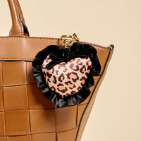 A brown woven handbag paired with The Darling Effect's Cheetah Love Puffy Heart Bag Charm—a heart-shaped keychain with cheetah print, black ruffle trim, and a small black bow—makes the perfect playful accessory.
