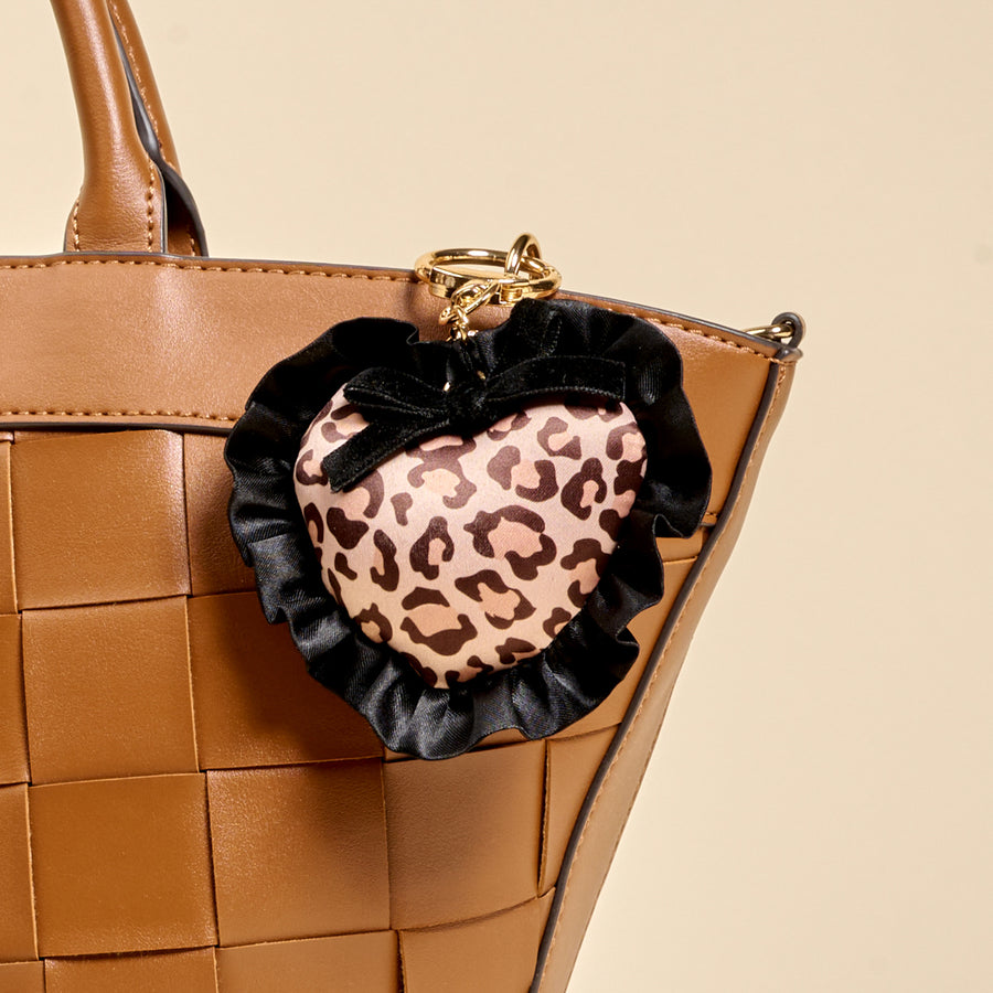 A brown woven handbag paired with The Darling Effect's Cheetah Love Puffy Heart Bag Charm—a heart-shaped keychain with cheetah print, black ruffle trim, and a small black bow—makes the perfect playful accessory.