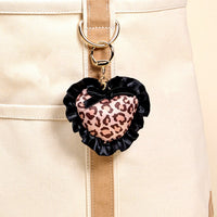 The Darling Effect’s Cheetah Love Puffy Heart Bag Charm features a leopard print, black ruffled edge, and small black bow, attached to a beige fabric bag with a gold clip for a stylish accessory.