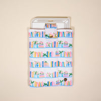 The Darling Effect Book Shelfie Book Cover: White fabric sleeve with a vibrant bookshelf print of pink, blue, and yellow books plus potted plants. Puffy polyester for protection. Gold snap flap. Packaged on a book-shaped card hanger.