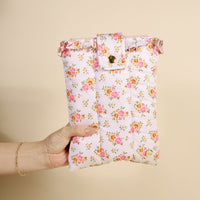 A hand holds the-darling-effect's Endless Daydream Cream Book Cover, a cute, puffy polyester cover with a pink and orange floral pattern and buttoned flap, set against a plain light beige background.