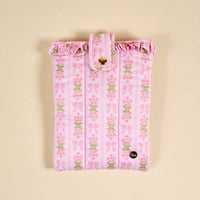 The Darling Effect’s Oh So Fancy Pink Book Cover is a puffy polyester pouch with a ruffled top, pink bows, and green floral designs. It has a buttoned strap closure and a small round label near the bottom.