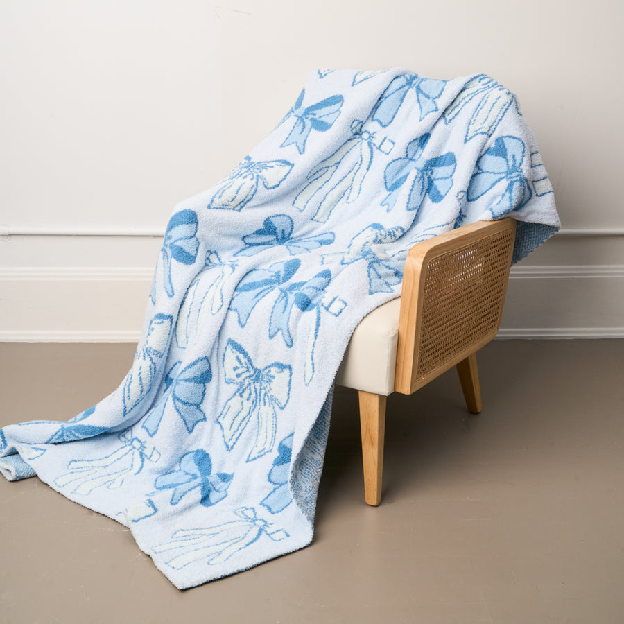 A Blushing Bows Blue Cozy Dreams Luxe Blanket by the-darling-effect, featuring dark blue bows, is draped over a modern wooden cane-back chair, set against a white wall and beige floor.