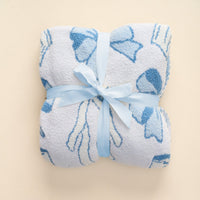 The-darling-effect’s Blushing Bows Blue Cozy Dreams Luxe Blanket features a plush, ultra-soft white fabric with blue bow patterns, neatly folded and tied with a light blue satin ribbon, set against a plain light beige background.