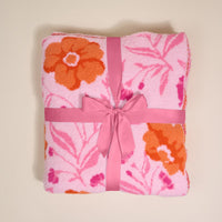 A folded Poppin' Petals Hot Pink Cozy Dreams Luxe Blanket by The Darling Effect, featuring orange and dark pink floral patterns and tied with a pink ribbon, is displayed on a light beige background for added charm and comfort.