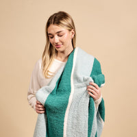 A young woman in a white sweater smiles while holding The Darling Effect Sicily Stripes Teal Cozy Dreams Luxe Blanket, featuring ultra-soft striped teal and light blue, against a beige background.