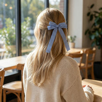 A woman with long blonde hair sits at a sunlit wooden table, facing away as she wears The Darling Effect's Gingham Blue Double Take Hair Bow with a cream sweater, showcasing her charming accessory indoors.