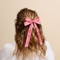 A person with wavy, light brown hair styled half-up wears The Darling Effect’s Gingham Red Double Take Hair Bow, shown from behind against a plain beige background.