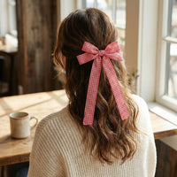 A person with wavy brown hair sits at a wooden table by a window, wearing a cream sweater and the Gingham Red Double Take Hair Bow from The Darling Effect. A beige mug in front of them adds to the cozy vibe of their chic hair accessory.