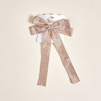 The Gingham Tan Double Take Hair Bow by The Darling Effect features a brown and white gingham pattern with long tails, displayed on a branded card—an adorable addition to your hair accessory collection.