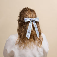 A person with wavy, shoulder-length hair seen from behind, wearing a white top and sporting The Darling Effect's Blue Heirloom Rosette Hair Bow—a striking accessory that adds charm to their hairstyle.