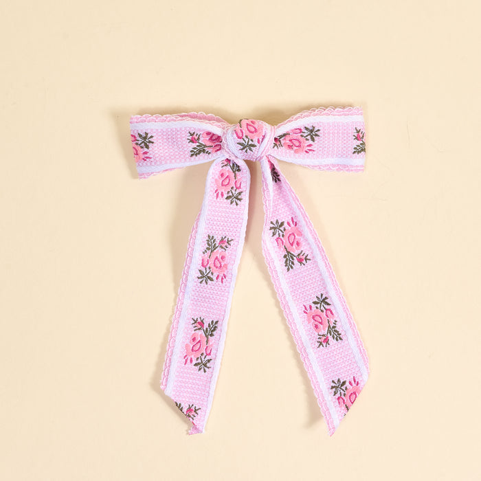The Darling Effect's Pink Heirloom Rosette Hair Bow features pink and white ribbon with embroidered flowers and green leaves, adding charm to your hair accessories collection. Shown on a neutral beige background.