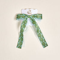 The Everbloom Forest Green Lovely Layered Hair Bow by The Darling Effect features long tails and comes on white packaging, ideal for stylish hair accessory lovers. Set against a light beige background.