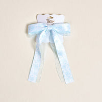 The Darling Effect's Something Blue Lovely Layered Hair Bow features a light blue hue with a white floral pattern and sheer ribbon tails, adding charm to your accessories. Shown on a cream background.