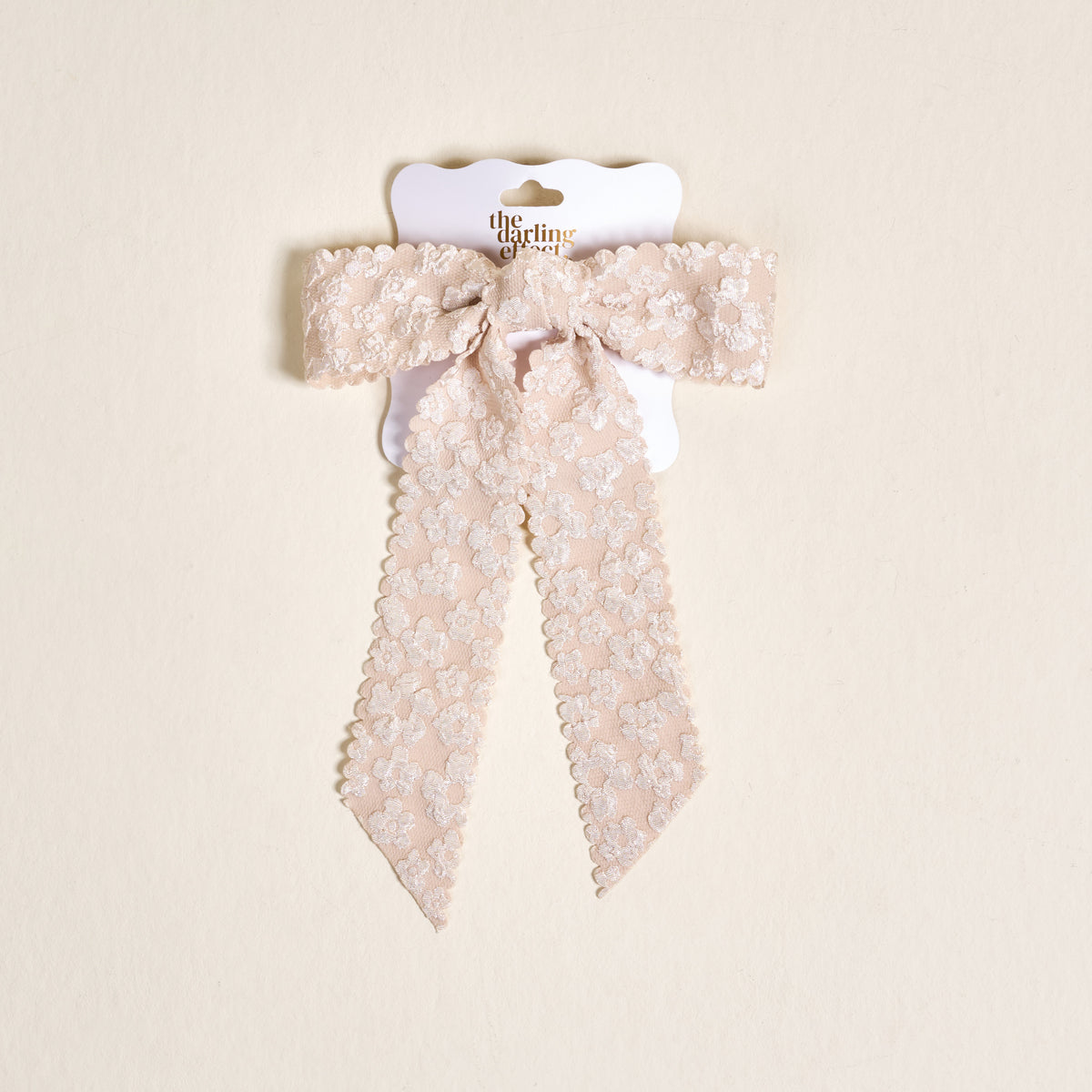 The Champagne Flower Scallop Hair Bow by the-darling-effect features pale pink lace with a floral pattern and scalloped edges, clipped to a white card with gold text on a beige background—an elegant addition to your hair accessory collection.