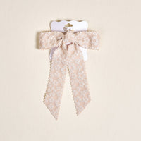 The Champagne Flower Scallop Hair Bow by the-darling-effect features pale pink lace with a floral pattern and scalloped edges, clipped to a white card with gold text on a beige background—an elegant addition to your hair accessory collection.