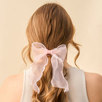 The Blush Sheer Pearl Hair Bow from the-darling-effect features a large, sheer pink bow adorned with white pearls and ribbon accents—a charming hair accessory for long, wavy blonde styles.